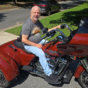 A smiling man sits on a shiny red trike motorcycle parked on a sunlit street. The background features green grass and trees, conveying a relaxed, joyful mood. Description:  Arc group home member on a Harley Davidson trike.