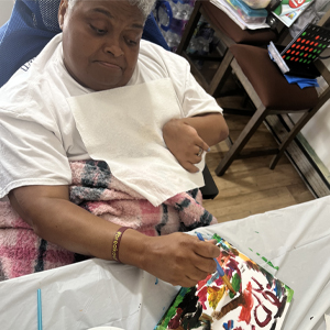 A person in a white shirt paints with vibrant colors on a small canvas, using a brush. A palette with various paints sits nearby on a table, conveying creativity. Description: A paint and sip party at a group home.