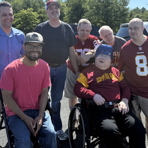 A group of seven men posing together in the Washington Commanders parking lot on a sunny day. Some are wearing sports jerseys, and one is in a wheelchair, smiling contently.  Description: Pre season fun at a Washington Commanders game.