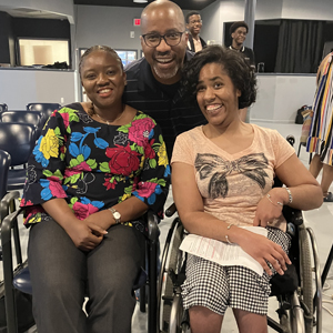 A joyful group photo shows two women sitting, one in a floral top, the other in a wheelchair with a beige shirt, and a smiling man standing behind them in a room. Description:  TEDx Largo rehearsal event at Creative Suitland.