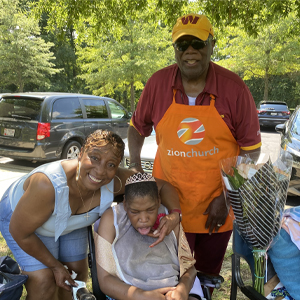 A smiling woman crouches beside a young person in a wheelchair, wearing a tiara. An older man stands nearby in an orange apron. Outdoors with parked cars.  Description: An outdoor birthday celebration.