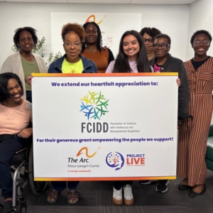A smiling woman crouches beside a young person in a wheelchair, wearing a tiara. An older man stands nearby in an orange apron. Outdoors with parked cars.  Description: An outdoor birthday celebration.
