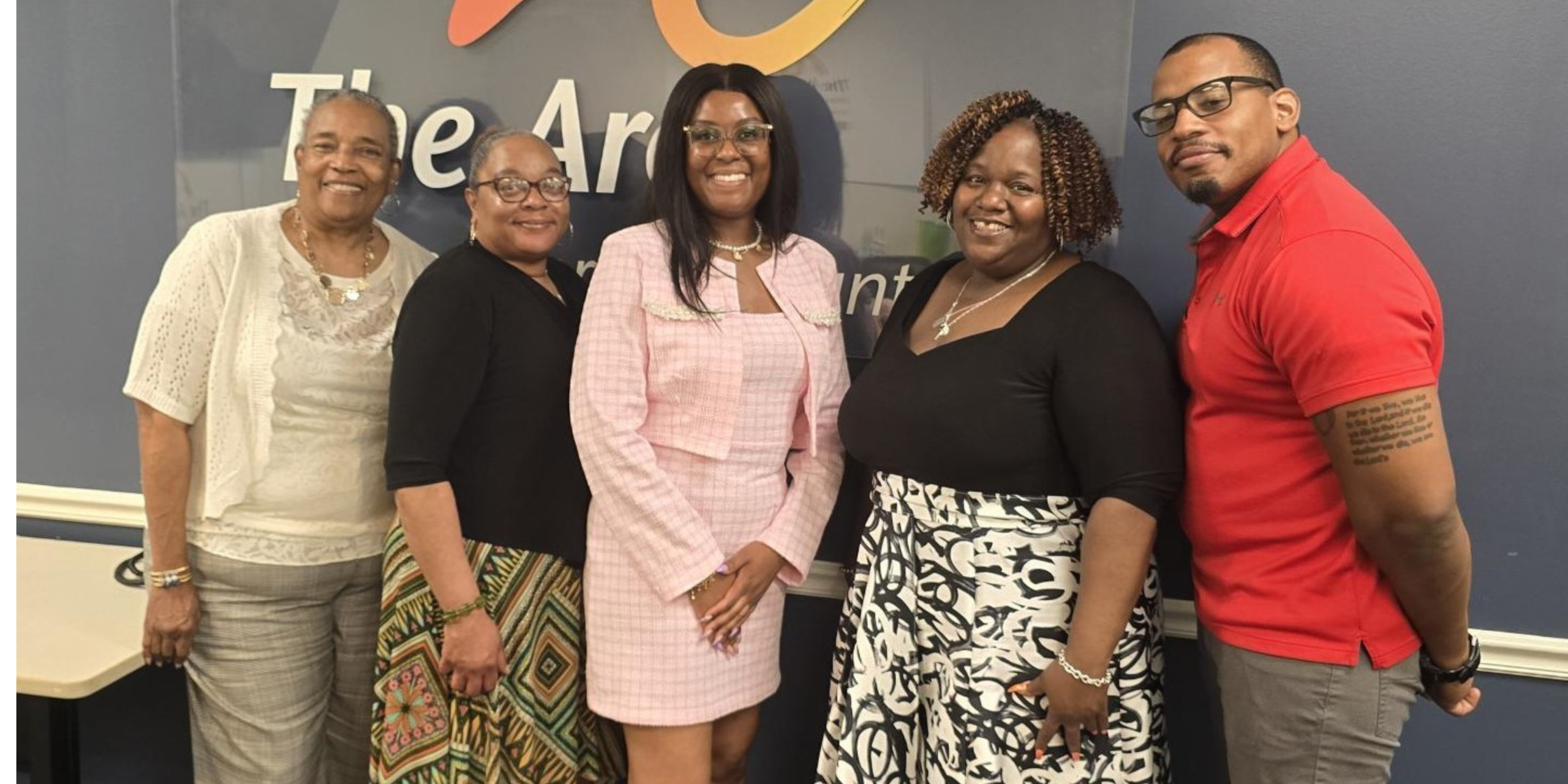 Five adults stand together indoors in front of a wall sign for The Arc, smiling at the camera.