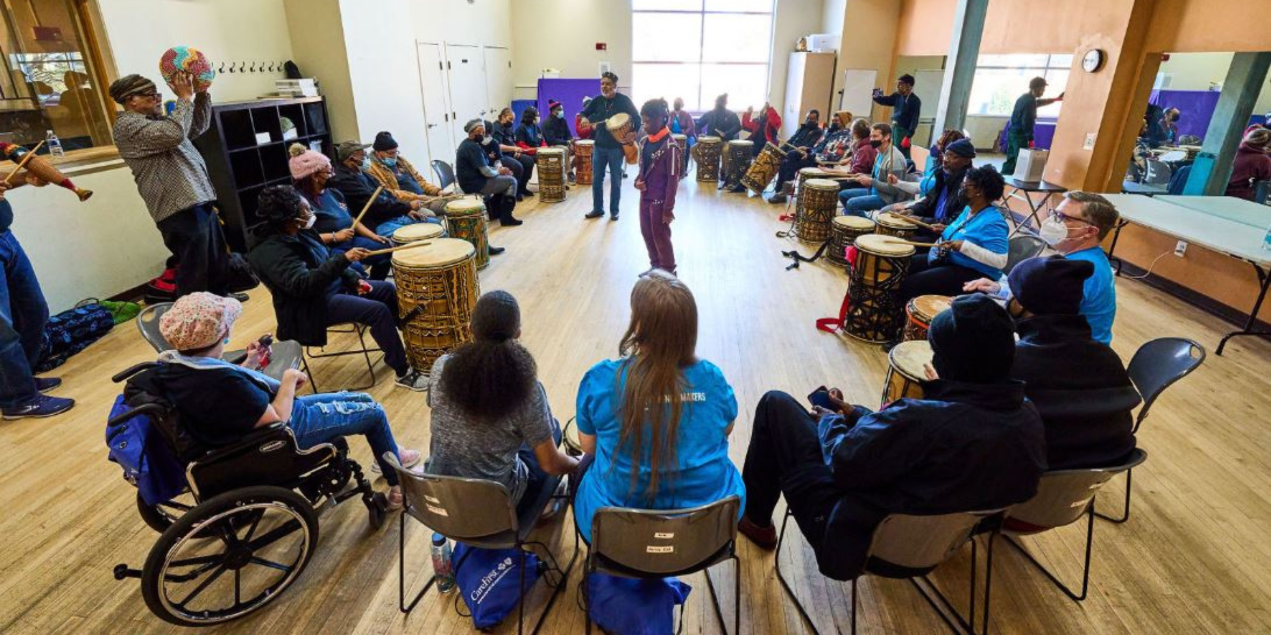 A diverse group of people sit in a circle playing hand drums together in a bright studio space, with a facilitator leading in the center.