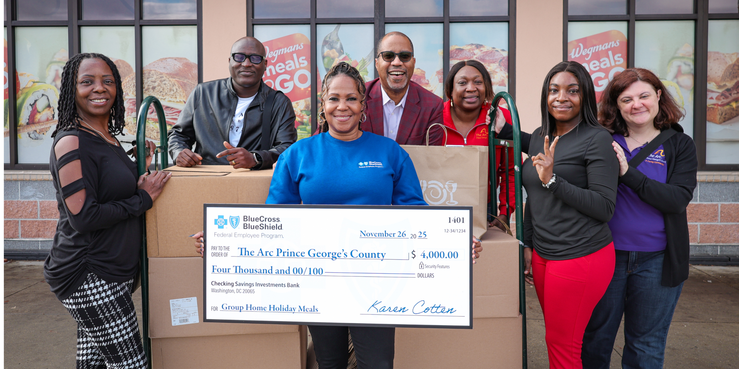 A group of seven adults stand outside a storefront holding a large ceremonial check made out to The Arc Prince George’s County for $4,000, surrounded by boxes of donated goods.
