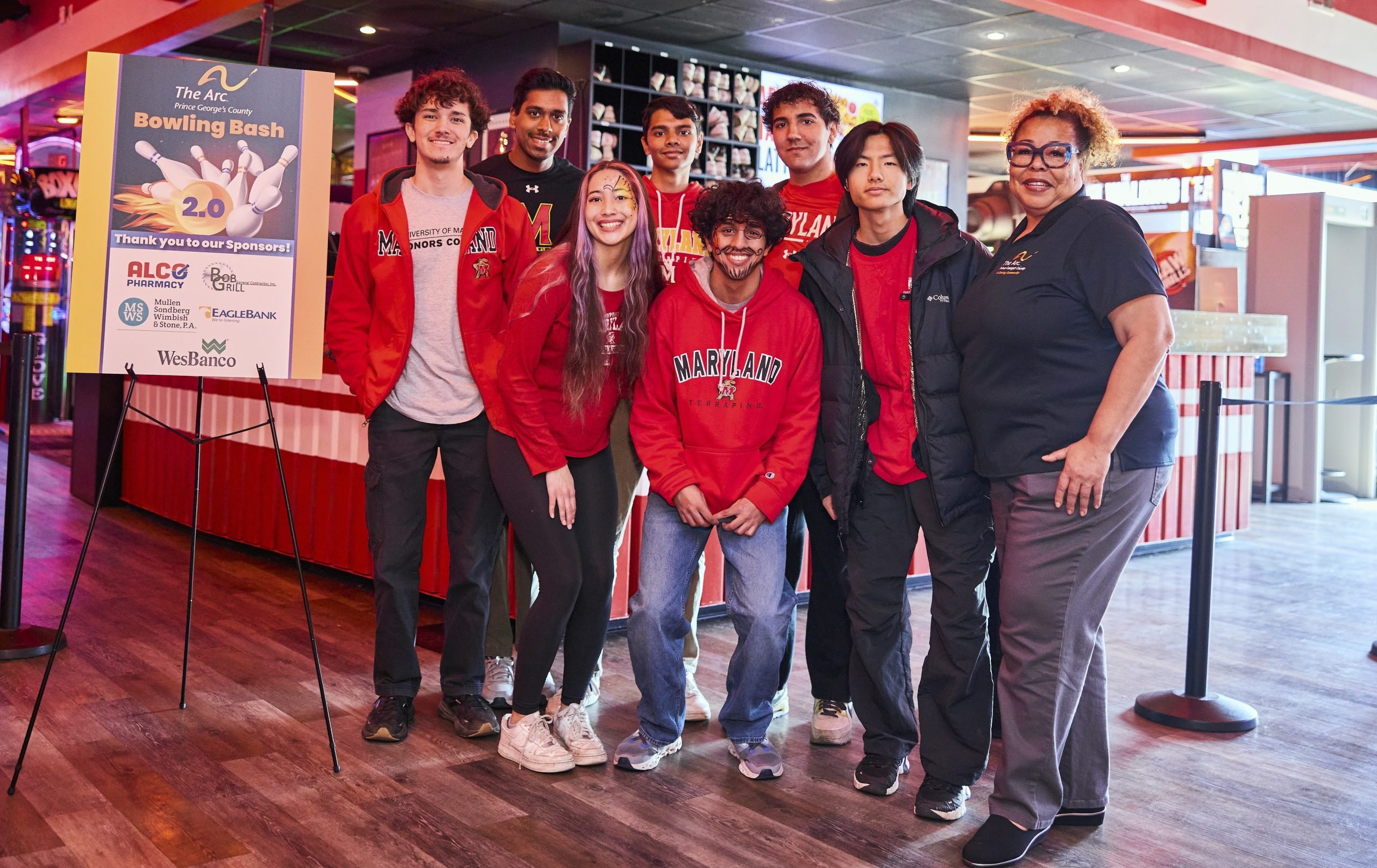 Group of eight people, wearing red and black, smiling and posing at a bowling alley. A sign reads "The ARC Bowling Bash 2.0." The atmosphere is cheerful.