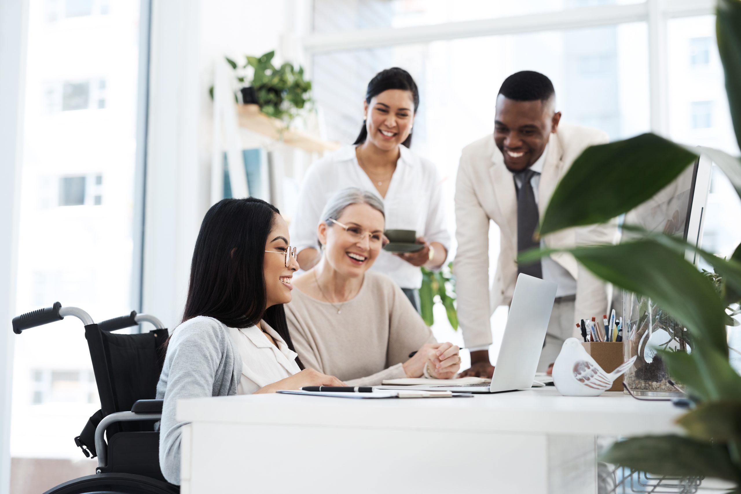 Diverse group of professionals, including a woman using a wheelchair, collaborating around a laptop during a meeting in a modern office setting.